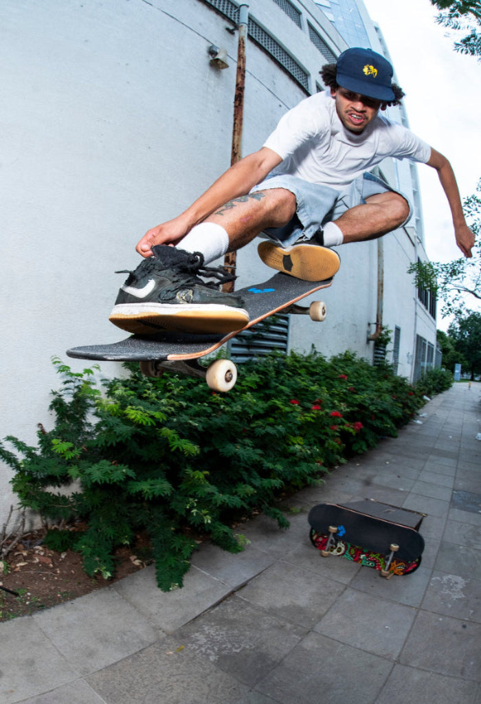 Pro skateboarder Bruno Vinicius performing a skateboard trick on a sidewalk with a building and greenery in the background. Flying on a 4Msb skateboard deck. Wearing a 4Msb cap dala horse navy with a yellow logo and dala horse on the front.