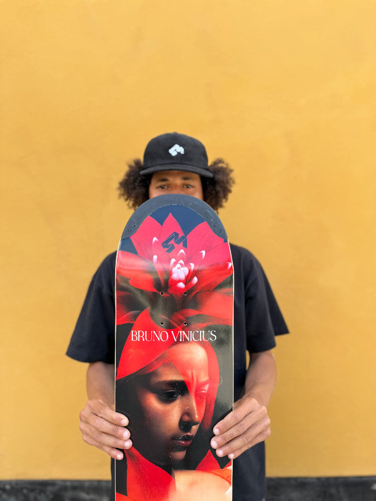 4Msb skateboard deck Bromeila with TIPtech, pro model for pro skateboarder Bruno Vinicius. The pro skater Bruno Vinicius is holding the deck in front of him, a yellow wall behind him. The graphic is red, black and white with a woman's face and the flower bromelia. Bruno is wearing a black 4Msb skateboard cap Quatro with a white 4Msb logo on the front. 