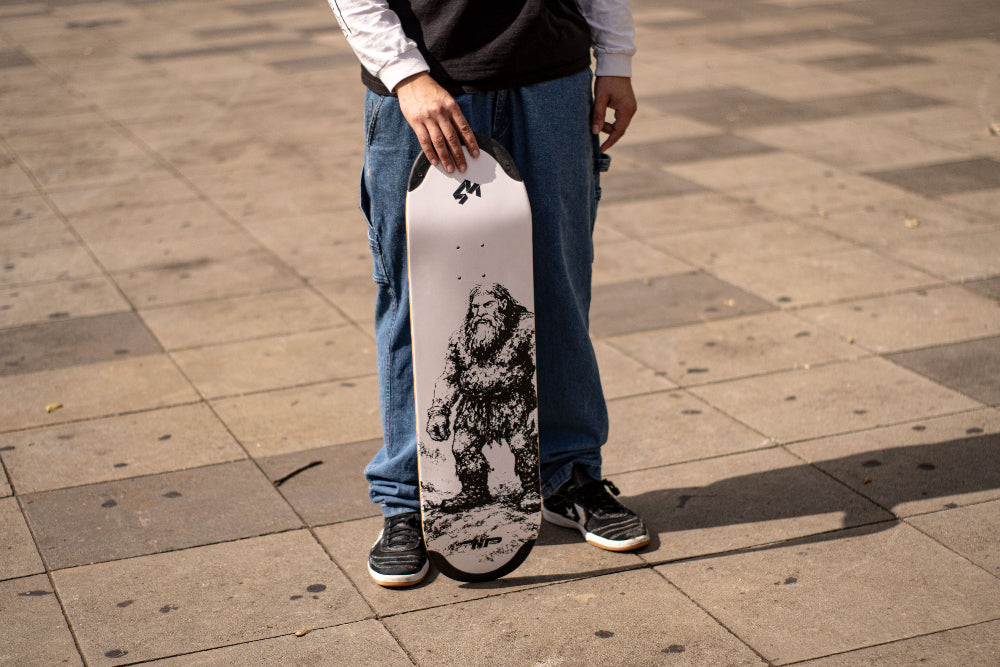 4Msb team rider Hadriel Junior, wearing jeans and black t-shirt, holding the new skateboard deck 4Msb Jättar (giants) with TIPtech in the streets of Sao Paulo, Brazil. The deck is black and white with a giant on a rocky ground, with white background. It's part of the folklore series.