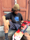4Msb pro skateboarder Bruno Vinicius with his pro model skateboard deck Bromeila with TIPtech. Sitting on a staircase with a red wooden door behind and the skateboard deck on his lap. The graphic is red, black and white with a woman's face and the flower bromelia. Bruno is wearing the 4Msb beanie Fisherman Edge in the colour biscuit.