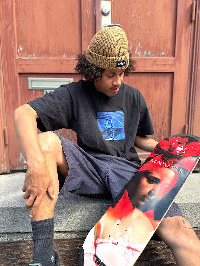 4Msb pro skateboarder Bruno Vinicius with his pro model skateboard deck Bromeila with TIPtech. Sitting on a staircase with a red wooden door behind and the skateboard deck on his lap. The graphic is red, black and white with a woman's face and the flower bromelia. Bruno is wearing the 4Msb beanie Fisherman Edge in the colour biscuit.