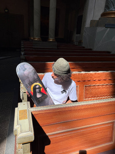 Pro skateboarder Bruno Vinicius holding his 4Msb pro model skateboard deck Saint Bridget, part of the saints collection, photographed inside the Oscar church in Stockholm, Sweden. The graphic is dark grey and has an image of Saint Bridget with a yellow halo and the title Bruno Vinicius.