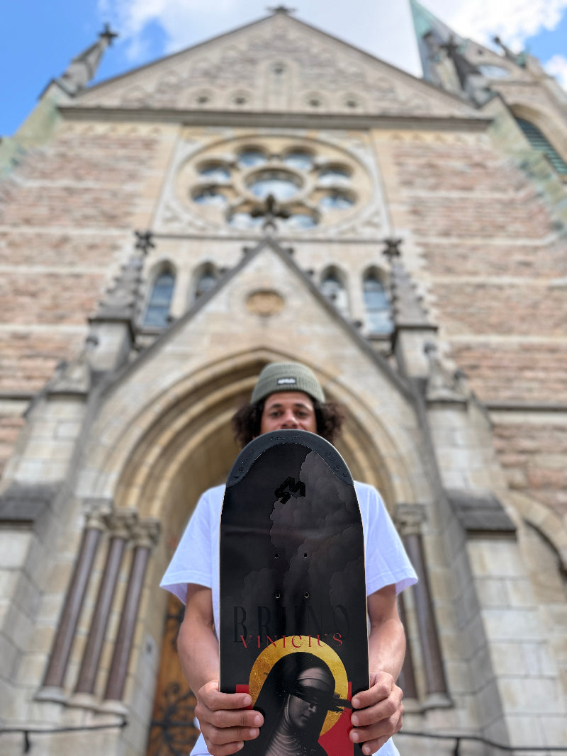 Pro Skateboarder Bruno Vinicius holding his 4Msb pro model skateboard deck Saint bridget, part of the saints collection, photographed outside the Oscar church in Stockholm, Sweden. The graphic is dark grey and has an image of Saint Bridget with a yellow halo and with the title Bruno Vinicius.