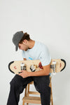 A skateboarder sitting on a wooden stool setting up a 4Msb skateboard deck Water Lily with TIPtech. He is wearing a grey 4Msb skateboard cap dala horse, an ice blue 4Msb t-shirt dala horse, dark jeans and light blue and white sneakers.