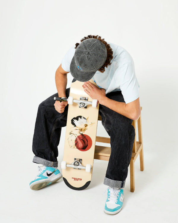 A skateboarder sitting on a wooden stool setting up a 4Msb skateboard deck Water Lily with TIPtech. He is wearing a grey 4Msb skateboard cap dala horse, an ice blue 4Msb t-shirt dala horse, dark jeans and light blue and white sneakers.