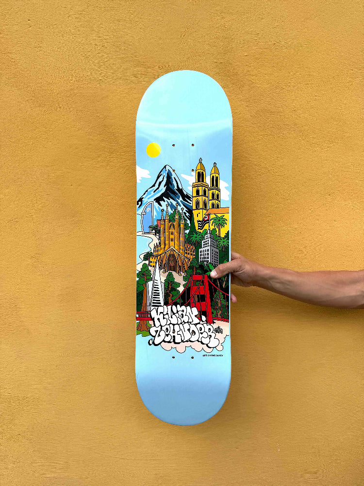 Pro skater Kilian Zehnder showing his new pro model, skateboard deck 'cities' classic shape (without TIPtech) against a yellow wall. The graphic of the deck has a light blue background, small yellow sun and skylines, mountains, towers, palm threes illustrating Kilian's four favorite cities.
