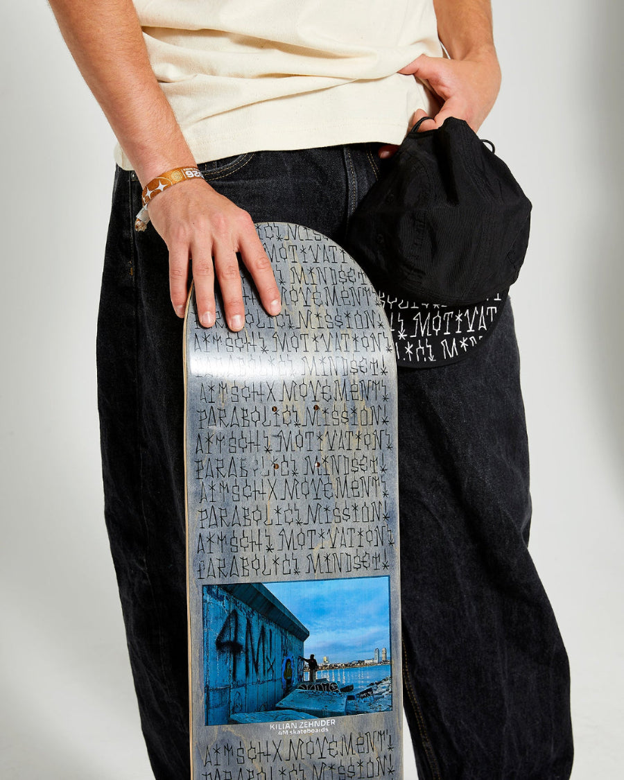 skateboarder holding a blue grey skateboard deck 4Msb graffiti Kilian Zehnder and a black cap graffiti Kilian Zehnder.