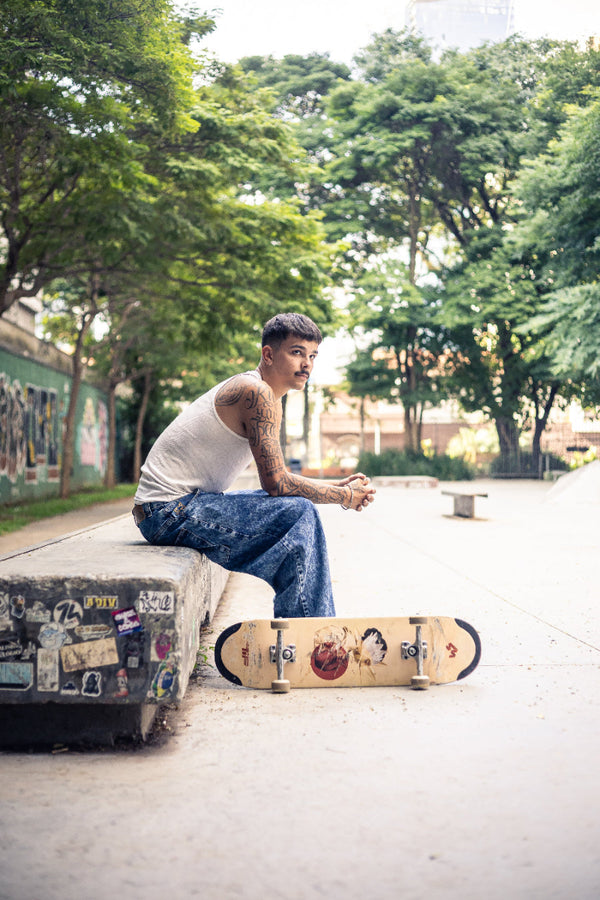 4Msb team rider Joao Richter, in baggy jeans and white tank top, sitting on a ledge with a 4Msb skateboard deck Water Lily TIPtech by his feet. Surrounded by green trees.