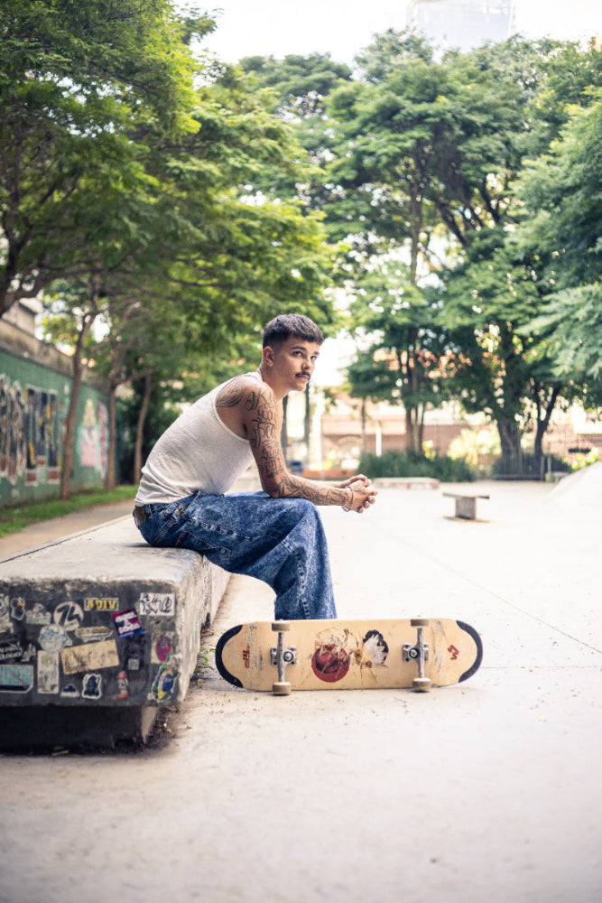 4Msb team rider Joao Richter, in baggy jeans and white tank top, sitting on a ledge covered by stickers, with a 4Msb skateboard deck Water Lily TIPtech by his feet, surrounded by green trees.