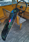4Msb skateboard deck Curupira, a man with hair on fire, on a dark background with the pro skateboarder name Bruno Vinicius in green. The board leans against a small wooden bench in an old bus.