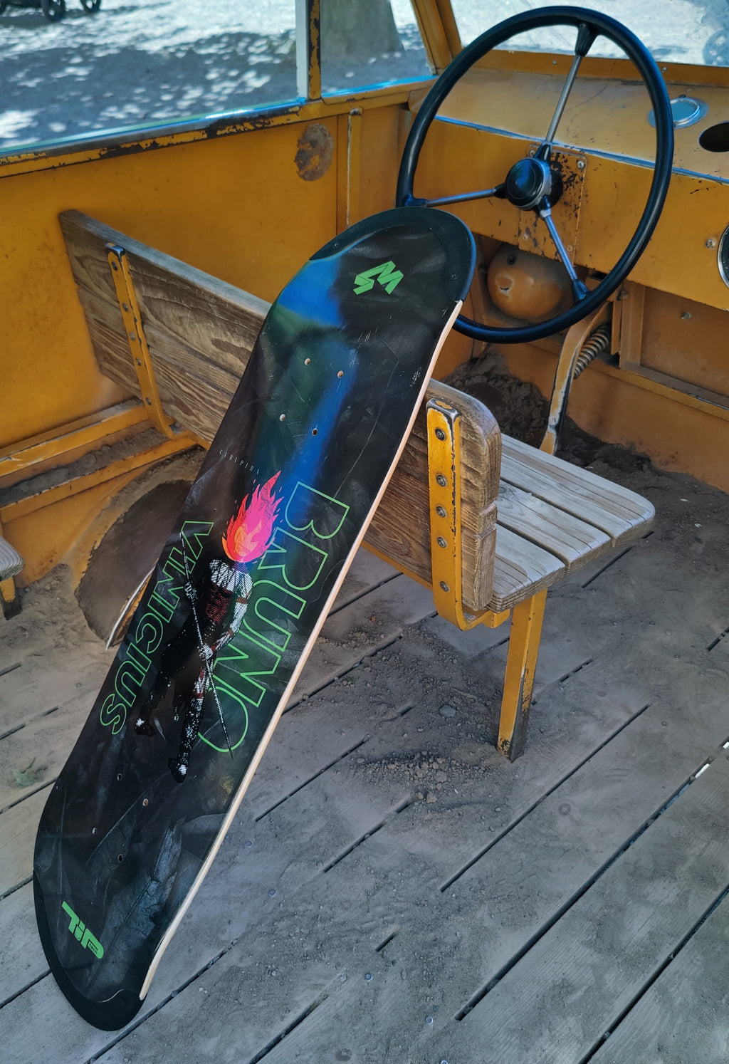 4Msb skateboard deck Curupira, a man with hair on fire, on a dark background with the pro skateboarder name Bruno Vinicius in green. The board leans against a small wooden bench in an old bus.