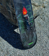 4Msb skateboard deck Curupira, a man with hair on fire, on a dark background with the pro skateboarder name Bruno Vinicius in green. The board stands in the gravel, leaning against a large grey stone.