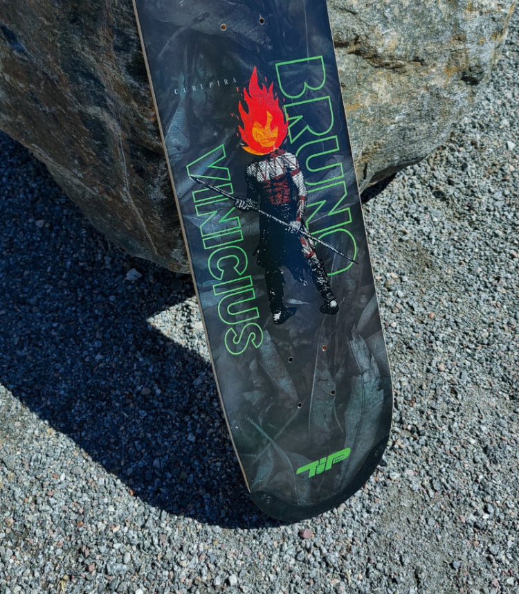 4Msb skateboard deck Curupira, a man with hair on fire, on a dark background with the pro skateboarder name Bruno Vinicius in green. The board stands in the gravel, leaning against a large grey stone.