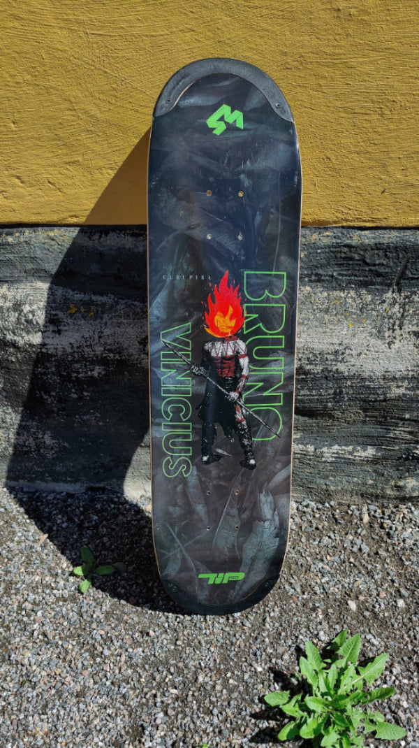 4Msb skateboard deck Curupira, a man with hair on fire, on a dark background with the pro skateboarder name Bruno Vinicius in green. The board stands in the gravel, leaning against a yellow wall.