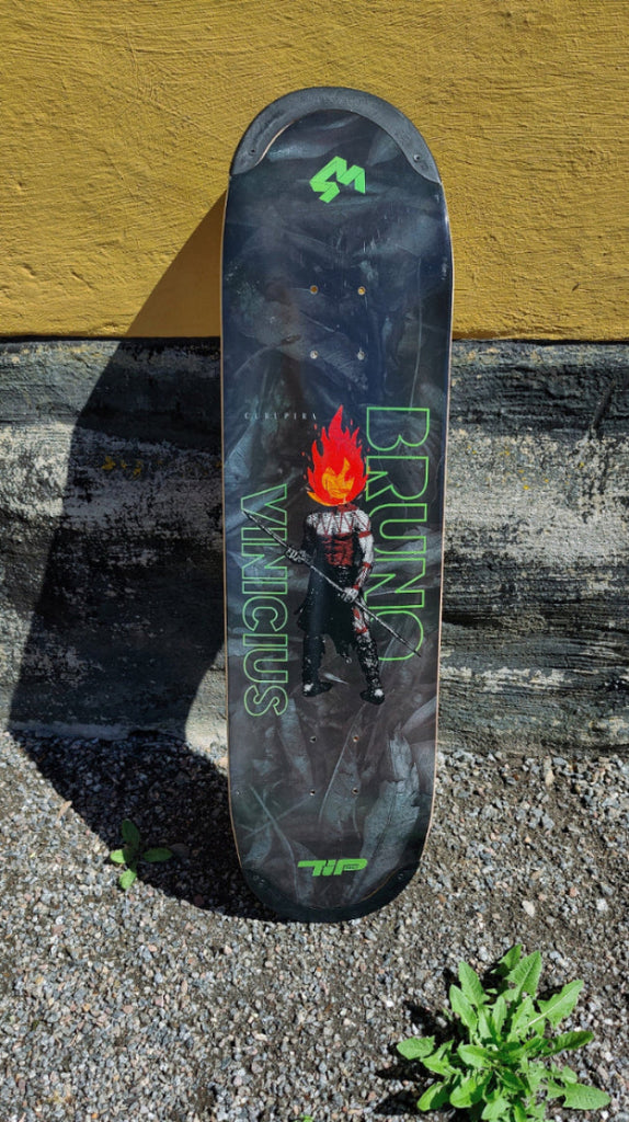 4Msb skateboard deck Curupira, a man with hair on fire, on a dark background with the pro skateboarder name Bruno Vinicius in green. The board stands in the gravel, leaning against a yellow wall.