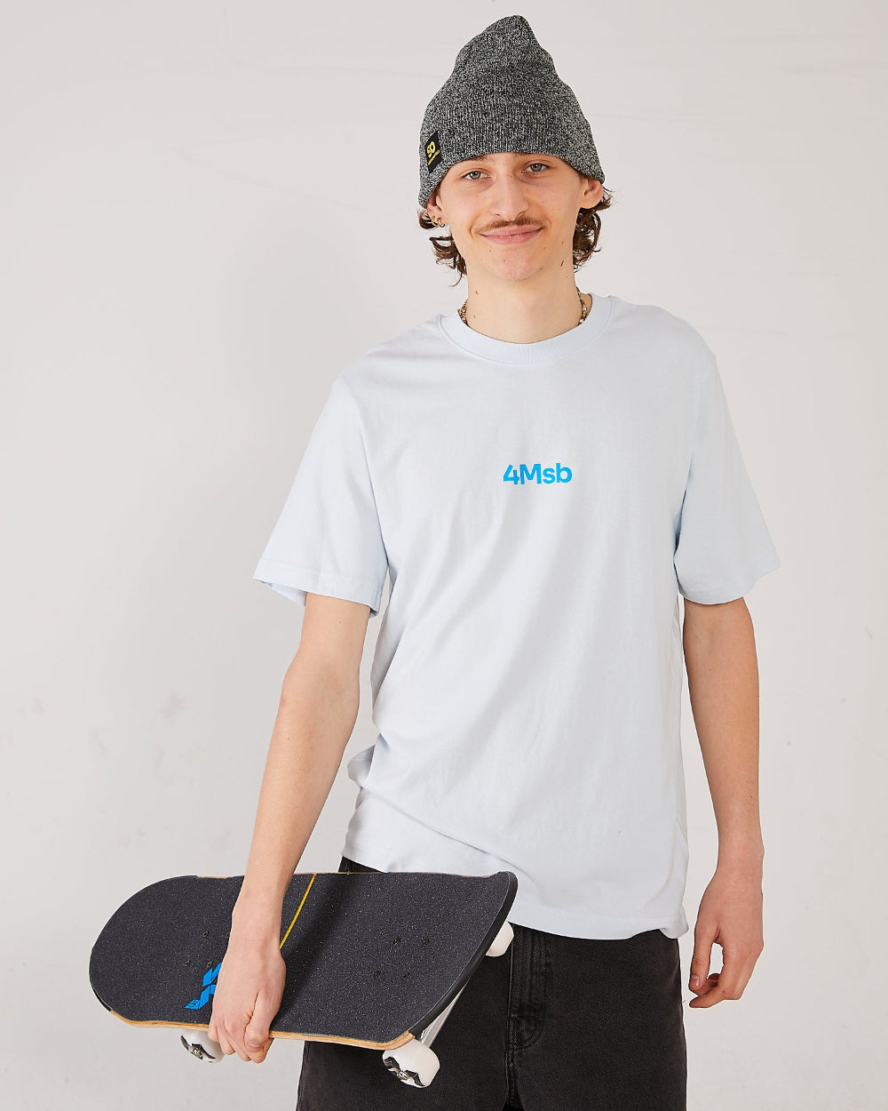 Skater wearing a light blue t-shirt with 4Msb logo, a antique grey cuffed beanie SO holding a skateboard.