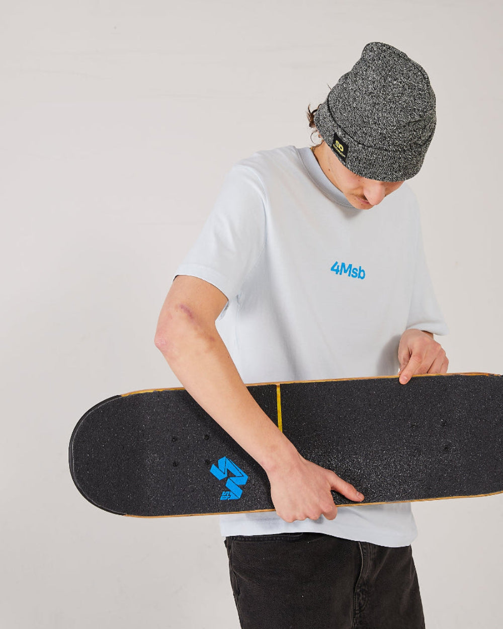 Skater wearing a ice blue 4Msb t-shirt, a antique grey SO cuffed beanie, holding a skateboard with a 4Msb griptape Quatro.
