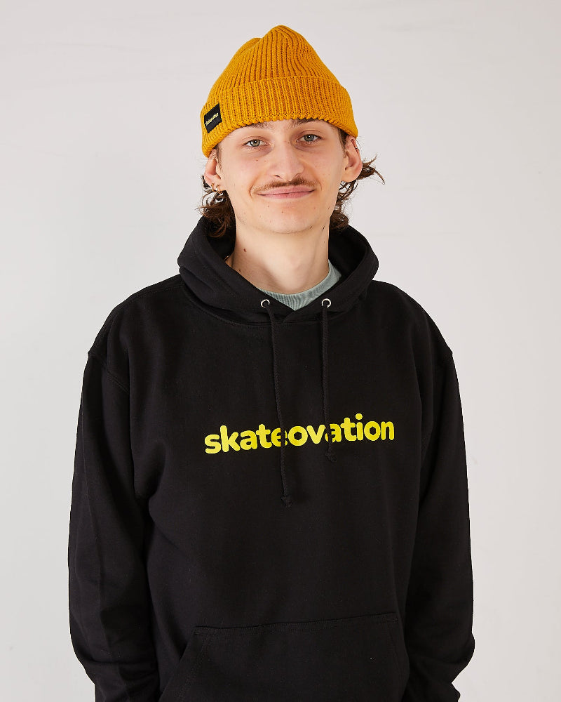 skater wearing a black hoodie with yellow 'skatevation' text and a mustard yellow harbour beanie.