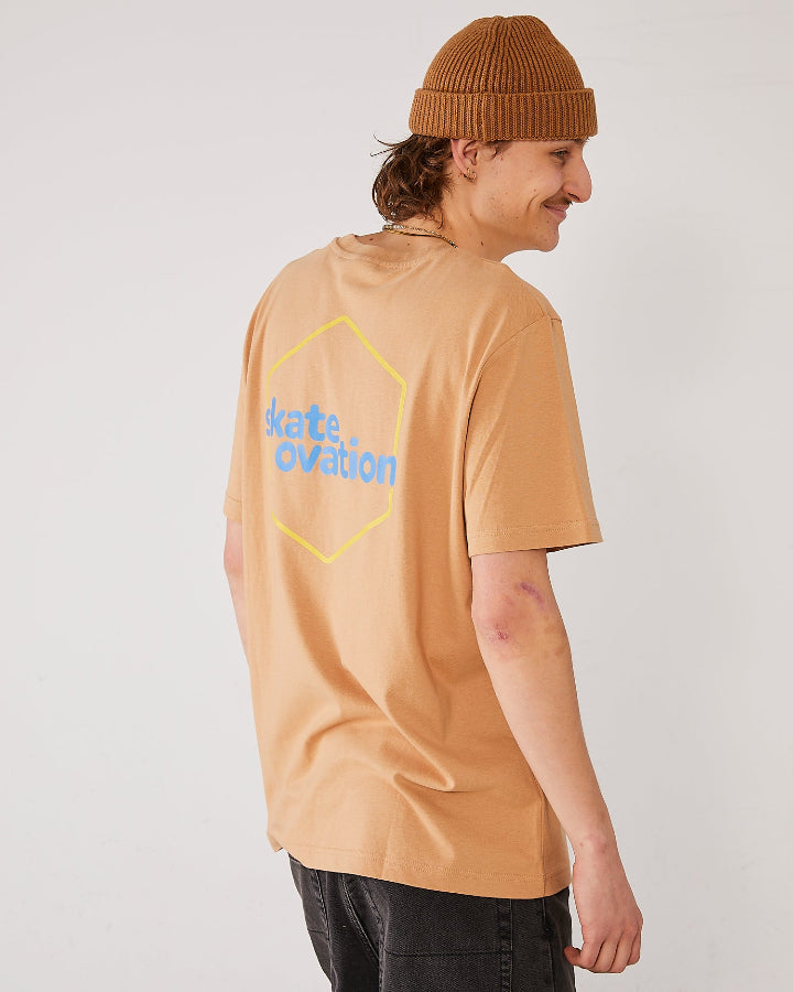 Skater seen from the side, his back facing the camera, wearing skateovation SO Honeycomb t-shirt in colour latte with a yellow honeycomb pattern on the back. skateovation written in blue at the center. Wearing a 4Msb fisherman beanie in the colour biscuit.