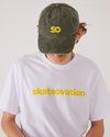 Skater facing forward, head down, wearing a Skateovation t-shirt core in white color with skateovation texted in yellow on the chest and a dad cap SO in the color vintage olive with SO embroidered in yellow on the front.