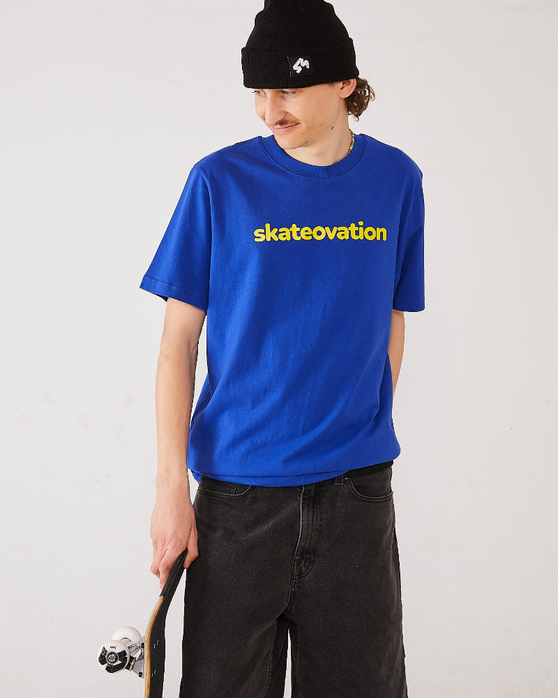 Skater wearing the Skateovation t-shirt Core in the color worker blue with skateovation texted in yellow on the chest, a 4Msb cuffed beanie Quatro in black and holding a TIPtech deck with 4Msb skateboard wheels Quatro in black and white.