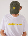 Skater facing forward wearing a SO dad cap in the color vintage olive and a white T-shirt with skateovation written in yellow on the chest.
