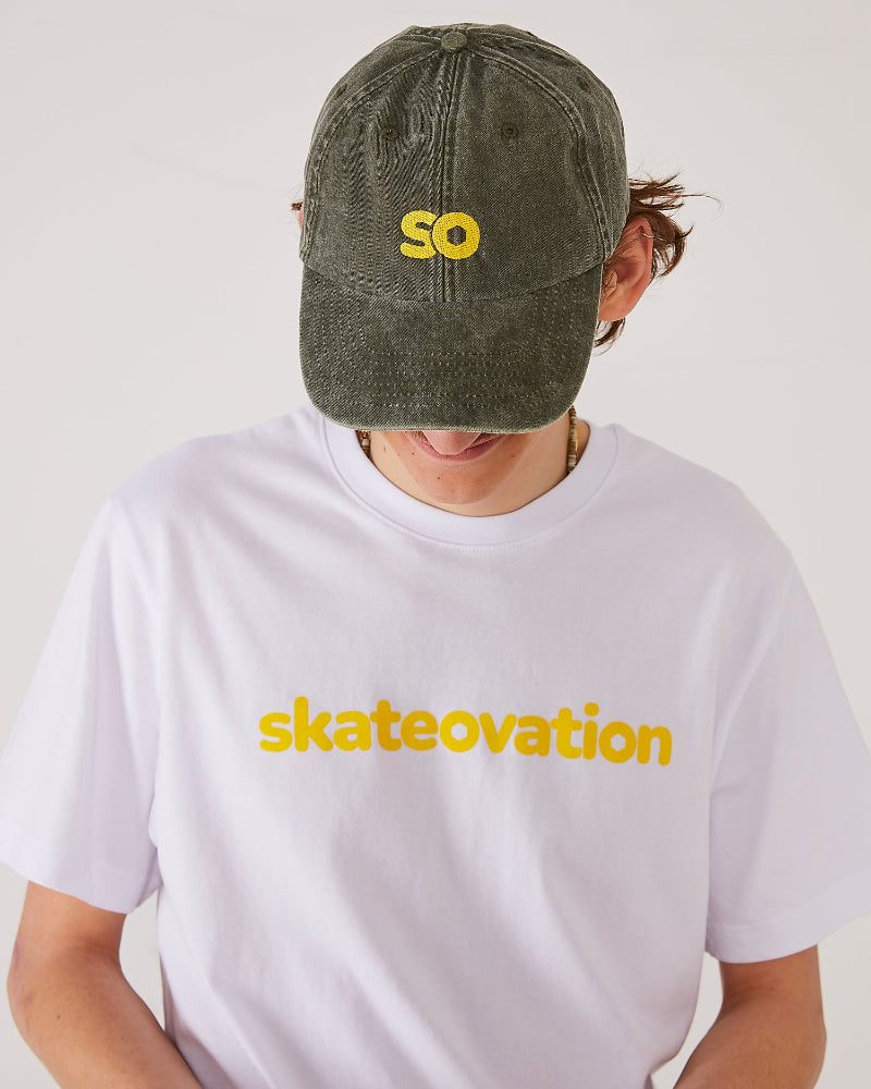 Skater facing forward wearing a SO dad cap in the color vintage olive and a white T-shirt with skateovation written in yellow on the chest.