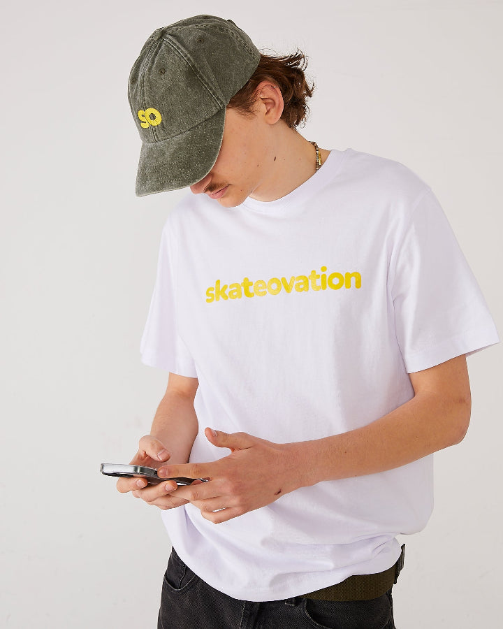 Skater facing forward looking at the mobile phone is his hands, wearing a skateovation SO dad cap in the color vintage olive and a white T-shirt with skateovation texted in yellow on the chest.